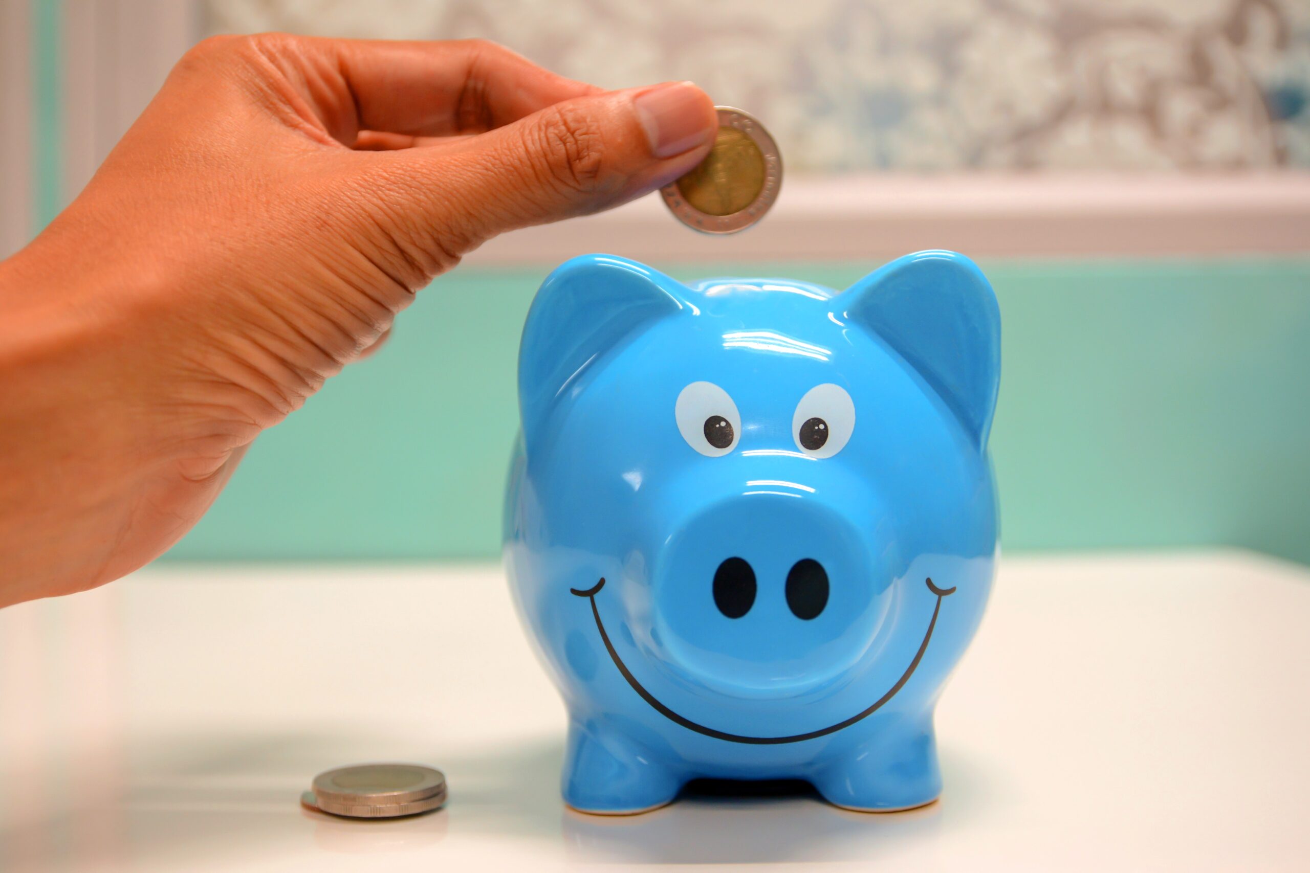 a blue piggy bank is getting a coin put in the slot on it's back. The piggy bank has a huge friendly smile